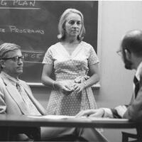 Nancy Fitzroy standing between two seated male colleagues at the GE Research Center. There is a chalkboard with writing on it behind them.  