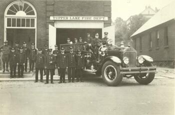 Group of firemen in uniform posing with fire truck out front of firehall at 21 High St.